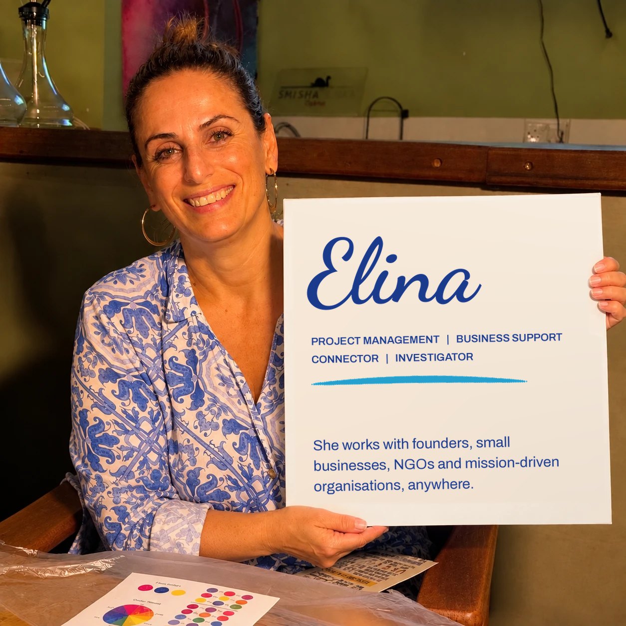 Elina smiling and holding a board that reads: Elina, Project Management, Business Support, Connector, Investigator. She works with founders, small businesses, NGOs and mission-driven organisations, anywhere.